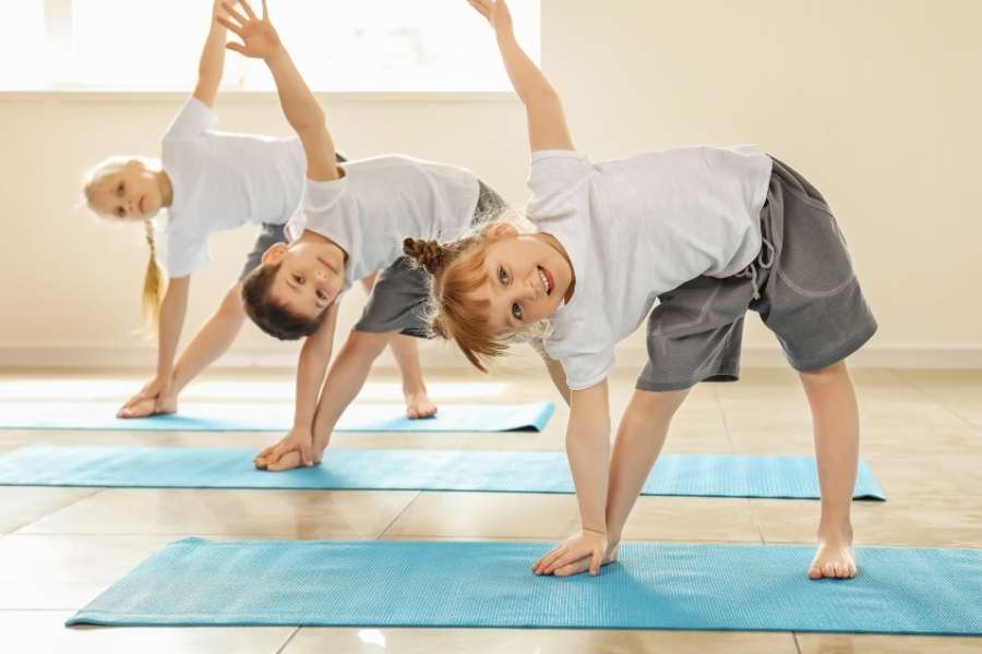 Trois enfants souriants en t-shirts blancs effectuent la posture du triangle lors d'une séance de yoga pour les enfants.