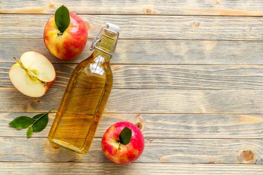 Une bouteille en verre remplie de vinaigre de cidre ambré est posée sur une table en bois rustique, entourée de pommes fraîches entières et coupées.